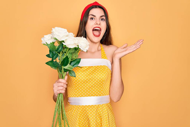 Молодая красивая пин-ап женщина в 50-е годы моды винтажное платье с бокетом цветов роз очень счастлив и взволнован, победитель выражение празднования победы кричать с большой улыбкой и поднятые руки
 - Фото, изображение