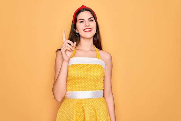 Молода красива жінка, одягнена в старовинну сукню 50 років на жовтому тлі, показує і вказує пальцем номер один, посміхаючись впевнено і щасливо
. - Фото, зображення