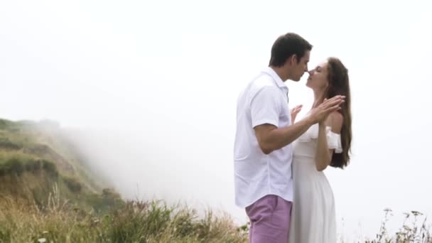 Young couple hugging on the edge of the cliff with thick fog on the background. Action. Tender man and woman enjoying each other above the clouds on a mountain. - Footage, Video