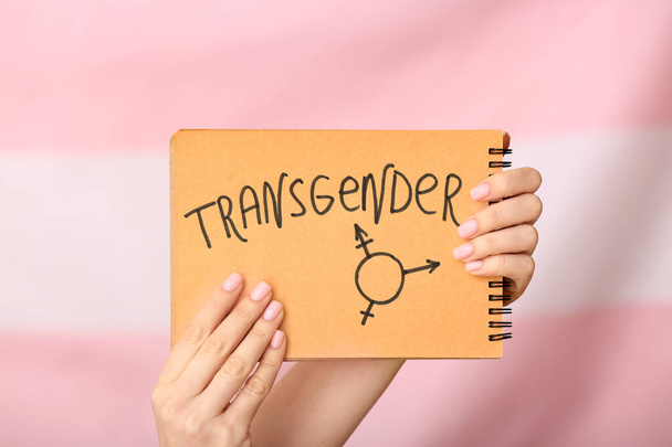 Female hands holding notebook with drawn symbol and word TRANSGENDER on color background - Photo, Image