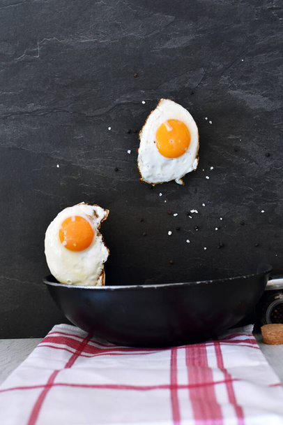 Play with perspective - fried eggs fly over a pan with recognizable egg white and yolk, salt, pepper and spices - concept for the classic, freshly fried egg as a healthy and simple meal - 写真・画像