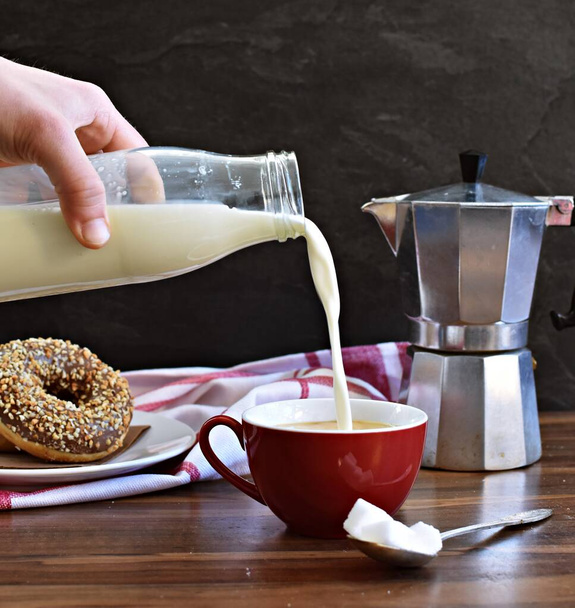 Verser le lait d'une bouteille en verre dans une tasse rouge pleine de café, qui est debout sur un fond en bois, avec un fond sombre et des beignets en arrière-plan
 - Photo, image