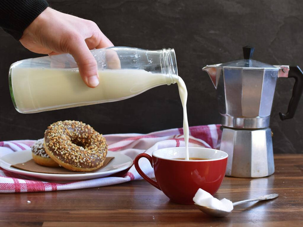 Налейте молоко из стеклянной бутылки в красную чашку, полную кофе, которая стоит на деревянном фоне, с темным фоном и пончиками на заднем плане
 - Фото, изображение