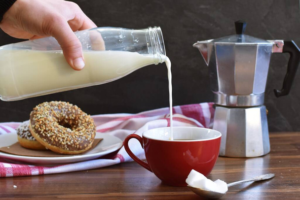 グラスボトルから牛乳を注ぎ、木製の背景に立っているコーヒーの完全な赤いカップに入れます。背景に暗い背景とドーナツ - 写真・画像