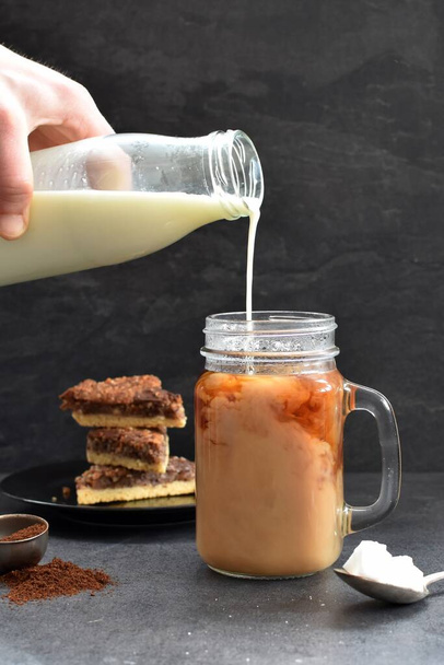 Налейте молоко в стакан полный кофе на темном фоне с ореховыми углами - свежеприготовленный горячий кофе против темной обстановки с молоком течет в нем
 - Фото, изображение