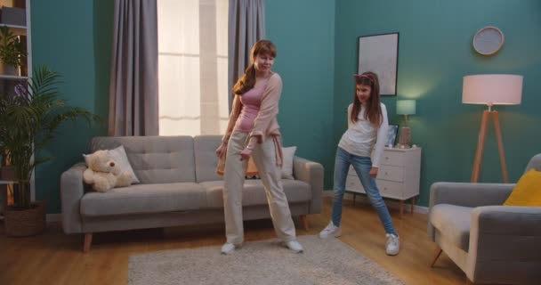 Cheerful pretty Caucasian mother showing dance moves to cute small daughter while they having fun together in living room. Woman and little teen girl dancing at home on weekend. Motherhood concept. - Filmati, video