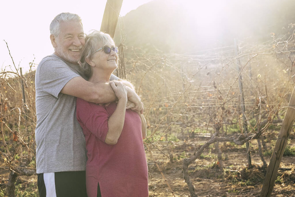 Cheerful couple of happy active senior man and woman couple - mature retired people in outdoor leisure activity in wine yard with sunlight in background  - Photo, Image