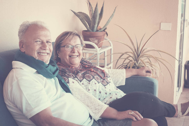 Χαρούμενο ηλικιωμένο ηλικιωμένο ευτυχισμένο ζευγάρι κάθεται στον καναπέ στο σπίτι χαμογελώντας και μένοντας μια χαρά κοιτάζοντας την κάμερα για μια οικογένεια πορτρέτων - Φωτογραφία, εικόνα