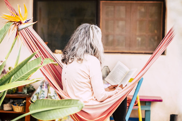 白い髪の多様性シニア女性は自宅で庭でハンモックでリラックス紙の本を読んで、一人で余暇時間の質を楽しむ - 写真・画像