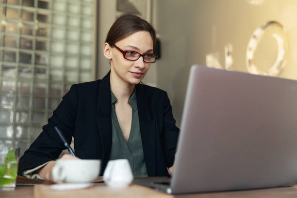 Focused business woman in stylish glasses sit on cafe working on laptop, concentrated serious female working with computer and cappuccino in coffee shop, freelancer, studying online, browse internet, checking bills - Фото, зображення