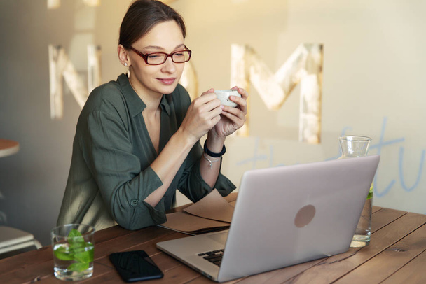 Woman in stylish glasses and black jacket drinking coffee at a restaurant and writing text on mobile laptop - Foto, Imagen