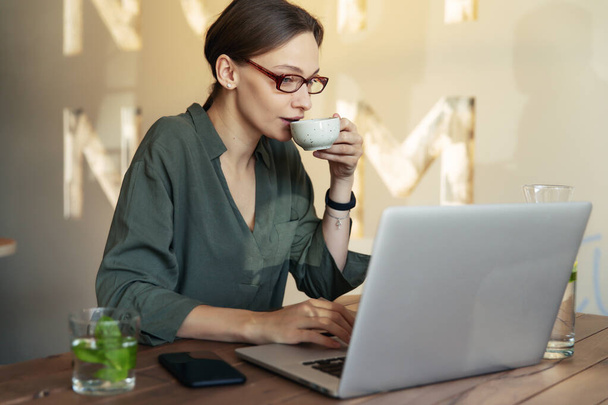 Woman in stylish glasses and black jacket drinking coffee at a restaurant and writing text on mobile laptop - Photo, Image