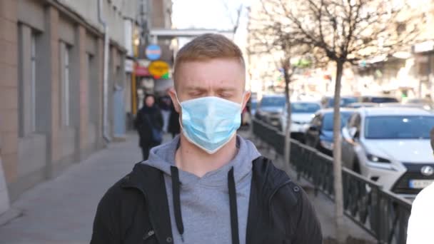 Dışarıdaki kalabalığın içinde virüsten koruyucu maske takan adam. Tıbbi maskeli genç adamın portresi şehir caddesinde duruyor. Coronavirüs salgınından sağIık ve güvenlik kavramı - Video, Çekim