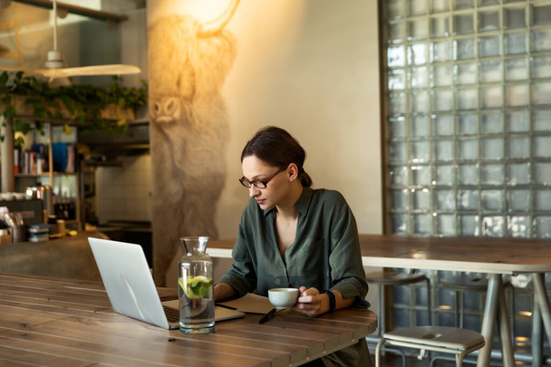 Woman in stylish glasses and black jacket drinking coffee at a restaurant and writing text on mobile laptop - Photo, image