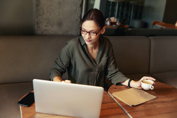 Business woman in a black suit and glasses sitting at a table in a cafe working at a laptop holding a notebook with records - Foto, Imagen