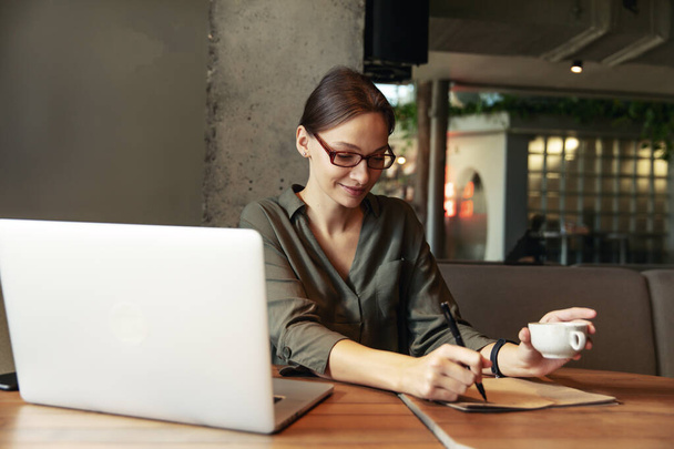 Business woman in a black suit and glasses sitting at a table in a cafe working at a laptop holding a notebook with records - Photo, image