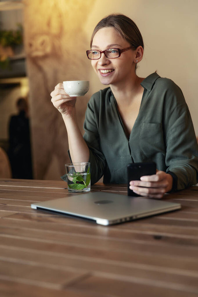 Cafe masasında oturan, dizüstü bilgisayarda çalışan şık gözlüklü, güler yüzlü iş kadını kameraya bakıyor. - Fotoğraf, Görsel