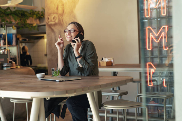 Attractive businesswoman in black jacket and stylish glasses sitting in cafe, talking on the phone and looking at laptop. - Foto, immagini