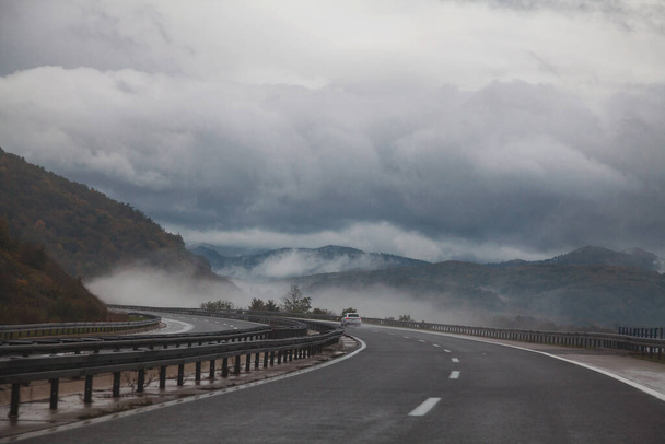 Šedá hladká asfaltová cesta v horách zatáčející vlevo, otevírající výhled na zamlžené hory za zamračeného dne - Fotografie, Obrázek
