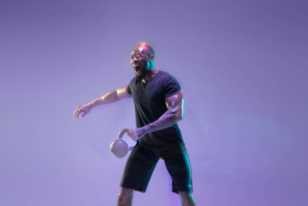 Young african-american bodybuilder training over purple background in neon, mixed light - 写真・画像