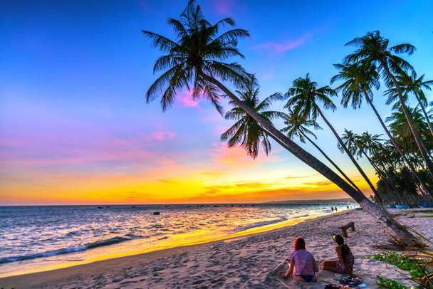 Закат на пляже с силуэтом пары, наклонными кокосовыми деревьями, длинными песчаными пляжами и красивым золотым небом и романтичным для курорта выходного дня
. - Фото, изображение