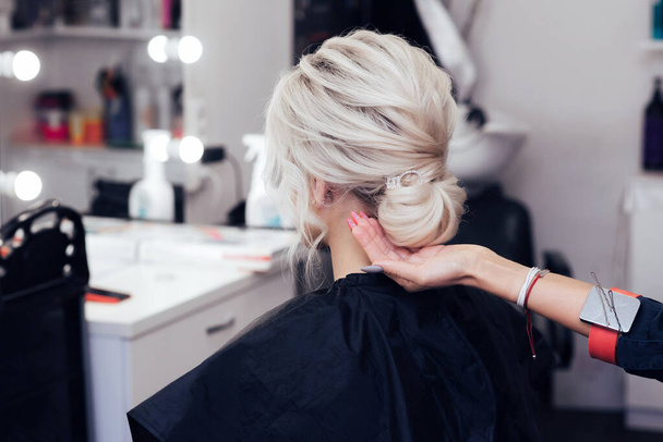 Friseurin macht ihre Haare zu einem Dutt auf dem Kopf eines blonden Mädchens mit langen Haaren - Foto, Bild