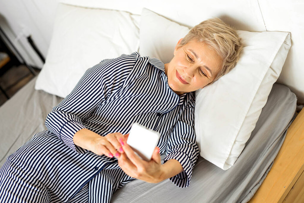  beautiful elderly woman in pajamas lies in bed with a phone - Fotó, kép