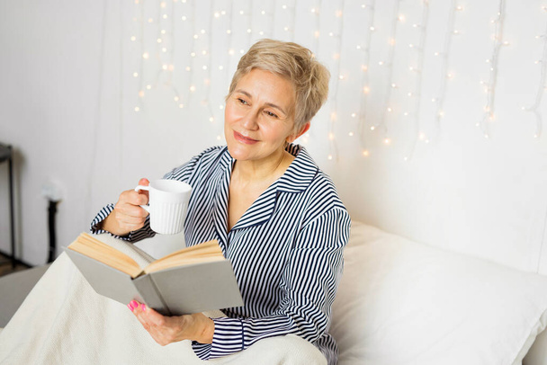  gyönyörű idős nő pizsamában ül az ágyban egy könyvvel és egy bögrével - Fotó, kép