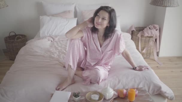 Portrait of happy Caucasian woman in nightwear dreaming as sitting on bed at home. Smiling brunette lady stretching in bedroom in the morning. Leisure, lifestyle, weekends. - Séquence, vidéo