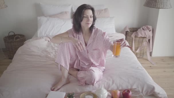 Happy charming Caucasian woman in pink pajamas taking glass with orange juice and drinking. Positive beautiful lady spending weekend morning in bedroom at home. Joy, leisure, happiness, lifestyle. - Felvétel, videó