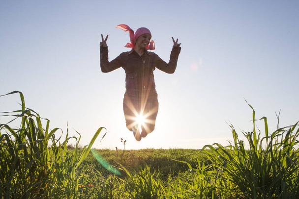 Молодая женщина в черной куртке и зеленых брюках прыгает с солнцем между ног на заднем плане, с розовым шарфом на голове и пальцами в победный жест на зеленом поле. Концепция рака молочной железы
. - Фото, изображение