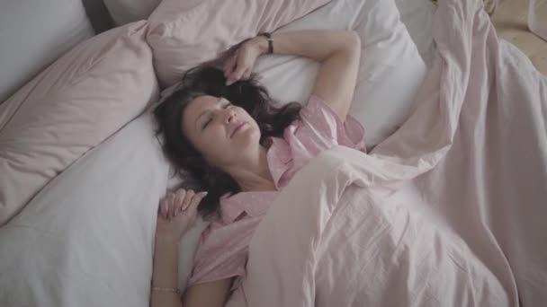Top view of positive adult Caucasian woman stretching in bed in the morning and smiling at camera. Portrait of happy brunette lady waking up in bedroom. Leisure, lifestyle, relaxation. - Кадры, видео