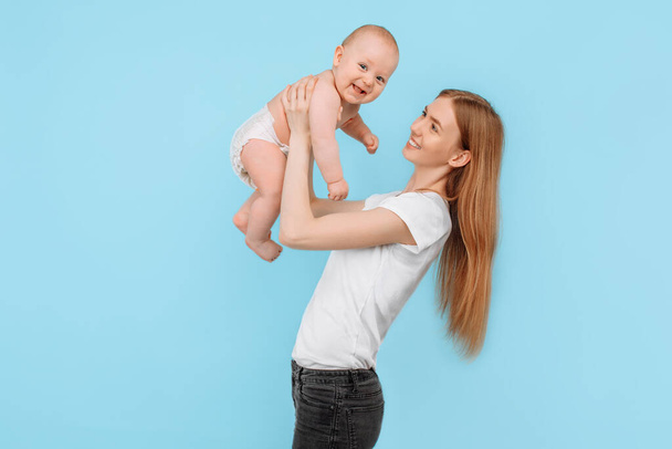 Família feliz. uma mãe rindo suavemente levanta seu adorável filho recém-nascido no ar, contra um fundo azul isolado
 - Foto, Imagem