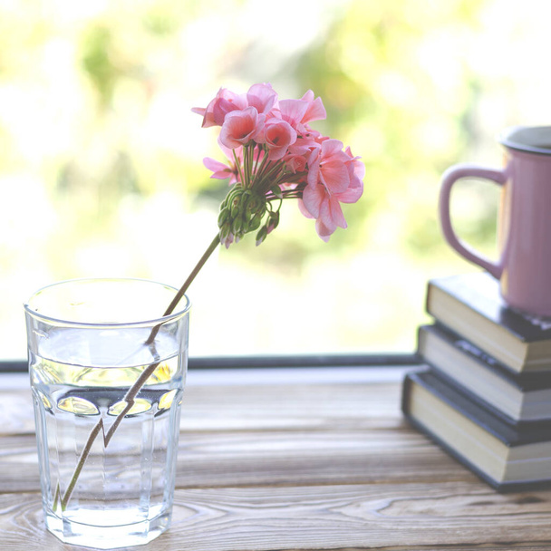Boeken zijn klaar om te lezen liggend op een houten vensterbank naast een glas met helder water, waarin een roze geranium bloem staat - Foto, afbeelding