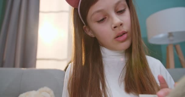Close up of Caucasian pretty small teen girl in funny ears reading book with fairytale and smiling. Cat on couch on background. Little child resting with faorytales at home. - Materiał filmowy, wideo