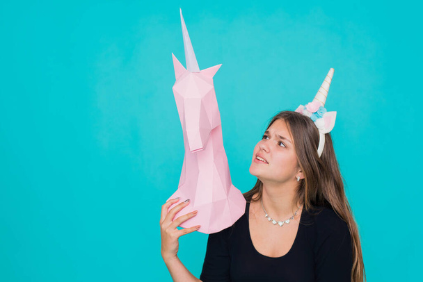 Kawaii teenage girl. Cute young woman with unicorn horn holding handmade paper pink unicorn over blue background. - 写真・画像