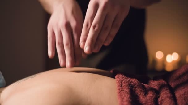 Jonge mannelijke massage therapeut doet vinger massage van een vrouw terug met een tatoeage in een massage kamer met schemerig licht op de achtergrond van kaarsen. Low-key premium massageconcept - Video
