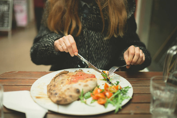 冬のコートを着た若い女性がアーケードの屋外のテーブルでカルゾーネピザを食べています - 写真・画像