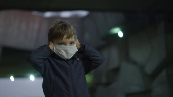 Boy in medical protective mask stands in shelter during pandemic outbreak of coronavirus COVID-19 and scared watches around. Kid closes ears because of loud emergency siren with red light outdoors.  - Materiał filmowy, wideo