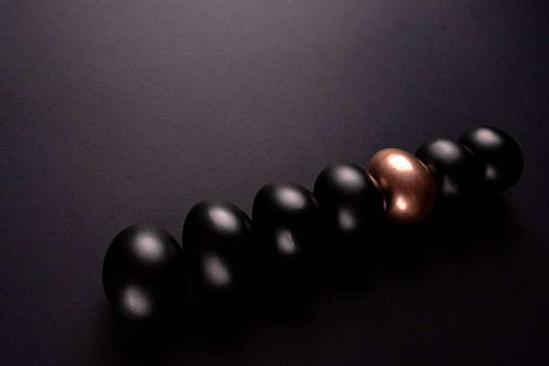 Ein goldenes Ei und schwarze Eier liegen hintereinander auf schwarzem Hintergrund mit Kopierraum - Foto, Bild