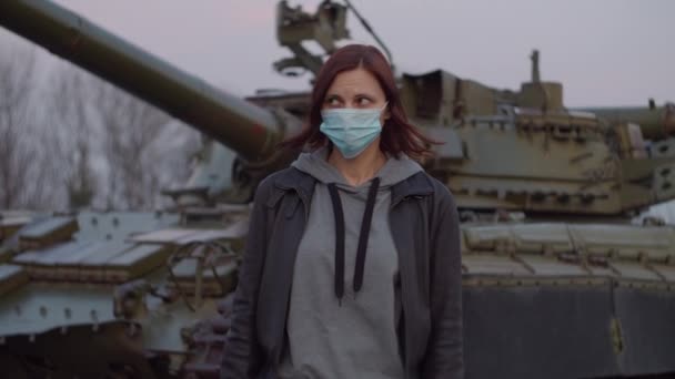 Young woman in medical protective mask stands by military machine during pandemic outbreak of coronavirus COVID-19 and scared looks around. Quarantine emergency and martial law to combat coronavirus.  - Footage, Video