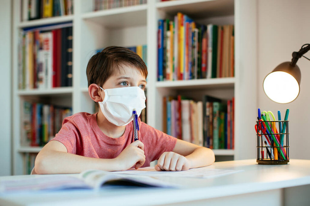 Παιδί που φοράει μάσκα προσώπου και μελετά στο σπίτι κατά τη διάρκεια της επιδημίας του κορωνοϊού. Νεαρός μαθητής που φοράει χειρουργική μάσκα και εργάζεται σε σχολικές εργασίες μόνος του. - Φωτογραφία, εικόνα