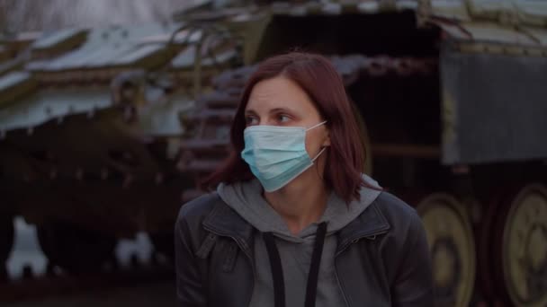 Woman in medical protective mask stands by military machine during pandemic outbreak of coronavirus COVID-19 and scared looks around. Quarantine and martial law to combat coronavirus. Close up - Séquence, vidéo