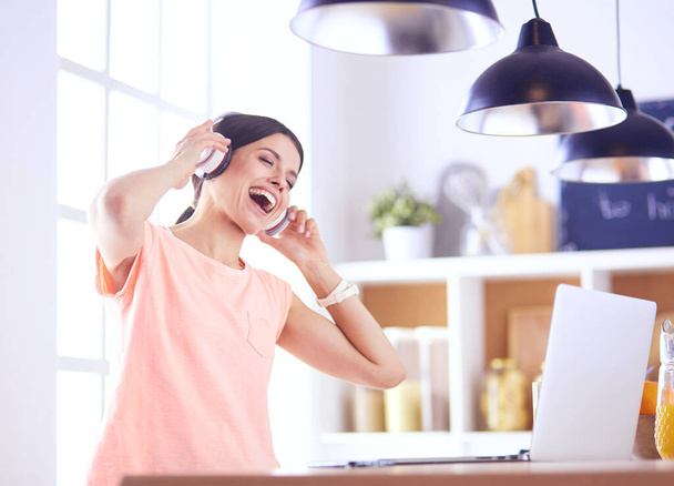 Portrait of a cheerful young woman listening to music with headphones and using laptop computer while standing at the kitchen - Фото, изображение