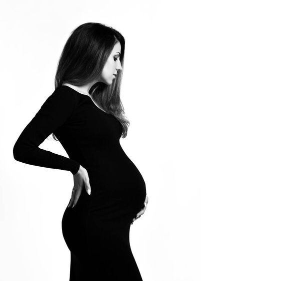 Black and white photo of pregnant female in tight dress. She put hands on her belly, posing isolated on white background. Close up - Fotó, kép