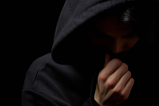  ein junger Kerl in einem schwarzen Kapuzenpullover auf schwarzem Hintergrund mit verdecktem Gesicht. - Foto, Bild