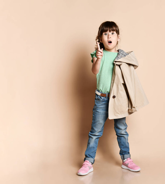 Cute girl posing standing in different poses, in jeans with belt, green t-shirt and beige coat and pink sneakers - Zdjęcie, obraz
