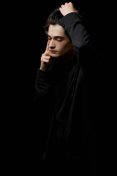  young guy in a black hoodie on a black background with a mobile phone in his hands - Foto, Bild