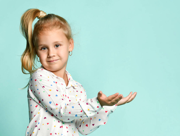 Little blonde girl with ponytail, in shirt with hearts print. She smiling, holding something posing on blue background. Close up - Fotoğraf, Görsel