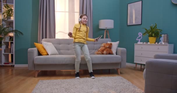 Caucasian teen small pretty girl in headphones listening to music and dancing in cozy living room. Cheerful cute kid dance and having fun at home. - Video, Çekim